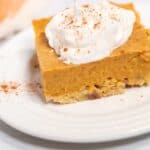 A square slice of Pumpkin Pie Dessert Bar with a crust base, topped with whipped cream and a sprinkle of cinnamon, served on a white plate.