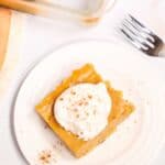 A square slice of Dessert topped with whipped cream and sprinkled with cinnamon sits on a white plate, with a fork and glass baking dish nearby.
