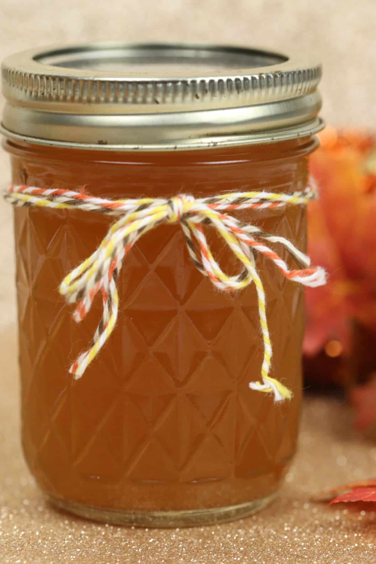 A glass jar filled with amber-colored Pumpkin Pie Moonshine, sealed with a metal lid and tied with a yellow and white string, sits on a neutral surface with autumn leaves in the background.