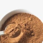 A close-up of ground cinnamon in a white bowl with a metal spoon scooping some of the spice, perfect as a Pumpkin Pie Spice substitute.