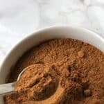 A white bowl filled with brown ground spice, ideal as a pumpkin pie spice substitute, with a metal spoon scooping some of the spice, on a white marble surface.