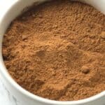 A white bowl filled with finely ground brown powder, perfect as a Pumpkin Pie Spice Substitute, placed on a white marble surface.