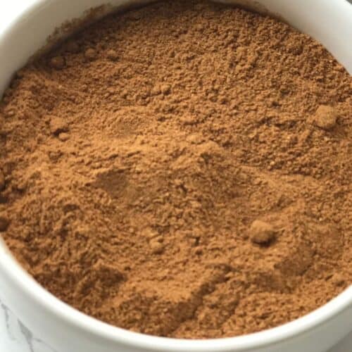A white bowl filled with fine, brown powder, perfect as a Pumpkin Pie Spice Substitute, sits on a white surface.
