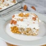 A slice of Pumpkin Poke Cake with white frosting and pecans on a white plate, with a pan of the same cake in the background.