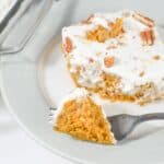 A slice of creamy Pumpkin Poke Cake topped with whipped topping and pecans sits on a plate, with a fork holding a bite in the foreground.