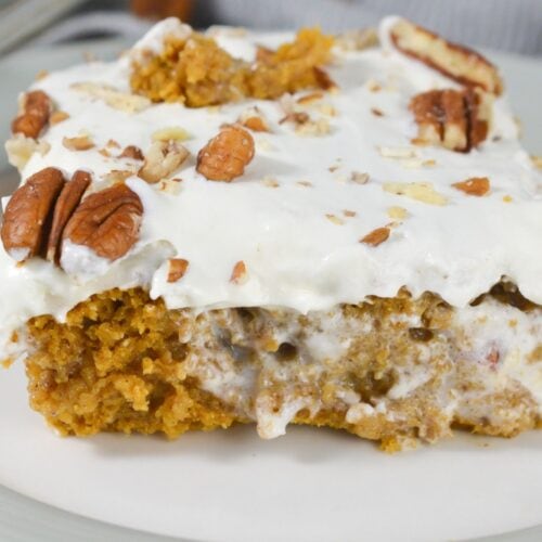 A slice of Pumpkin Poke Cake topped with white frosting and garnished with chopped and whole pecans sits beautifully on a white plate.