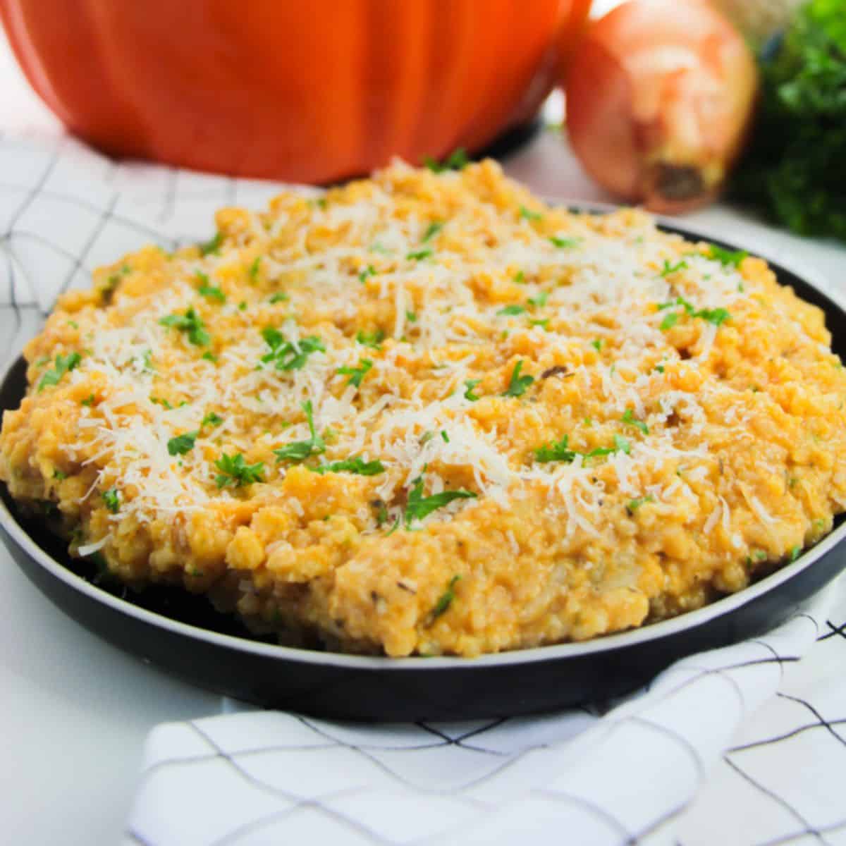 A plate of creamy pumpkin risotto topped with grated cheese and chopped herbs, set on a white surface with an onion and a pumpkin in the background.