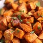 A white bowl filled with Brown Sugar Sweet Potatoes, roasted into golden cubes and garnished with chopped herbs.