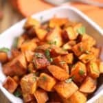 A white bowl filled with Brown Sugar Sweet Potatoes, garnished with chopped herbs, sits on a wooden surface with a spoon and orange napkin in the background.