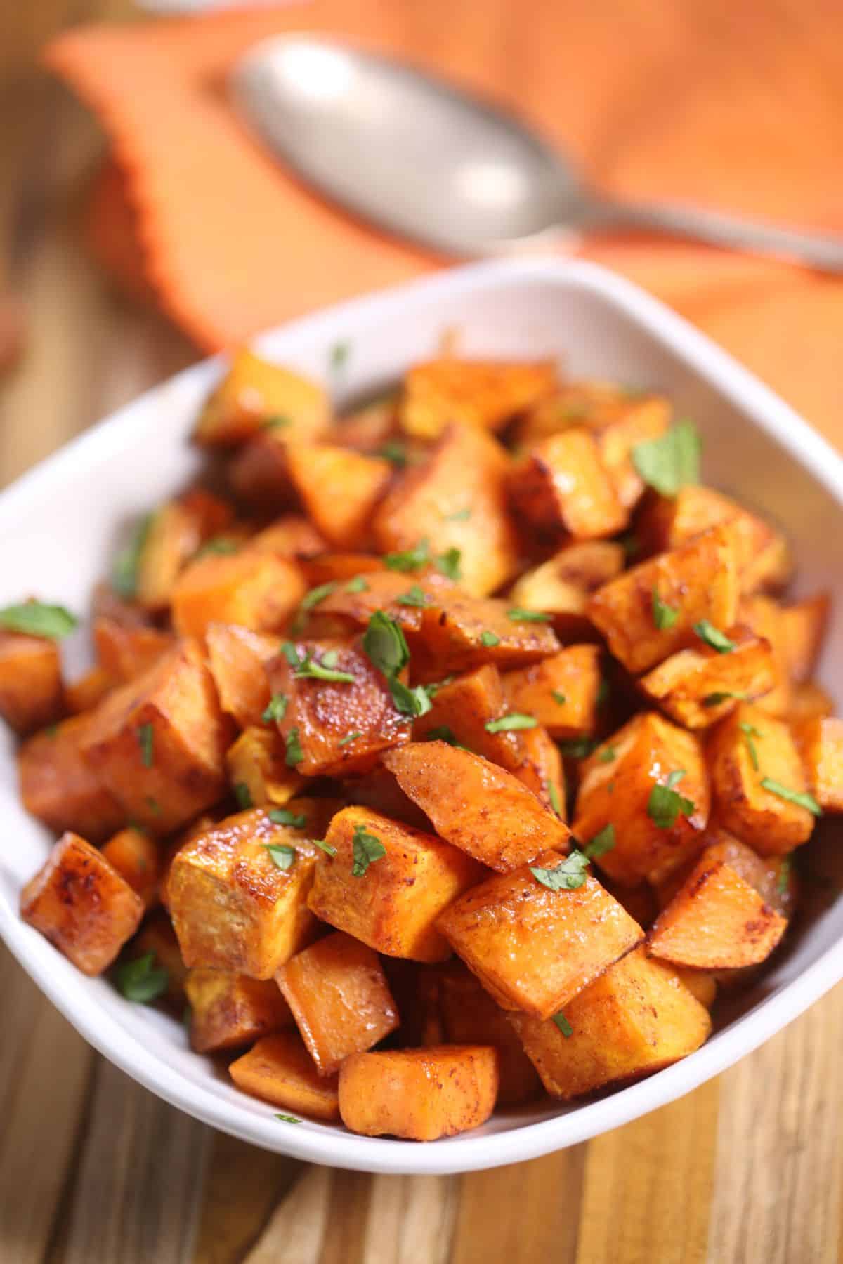 A white bowl filled with Brown Sugar Sweet Potatoes, roasted into golden cubes and garnished with chopped herbs, sits on a wooden surface with an orange napkin and spoon in the background.