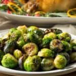 A plate of Roasted Brussels Sprouts with Parmesan Cheese sits on a wooden table, with a cooked poultry dish and garnishes visible in the background.
