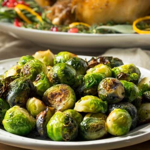 A plate of Roasted Brussels Sprouts with Parmesan Cheese sits in the foreground, with a cooked whole turkey garnished with herbs and citrus slices in the background.
