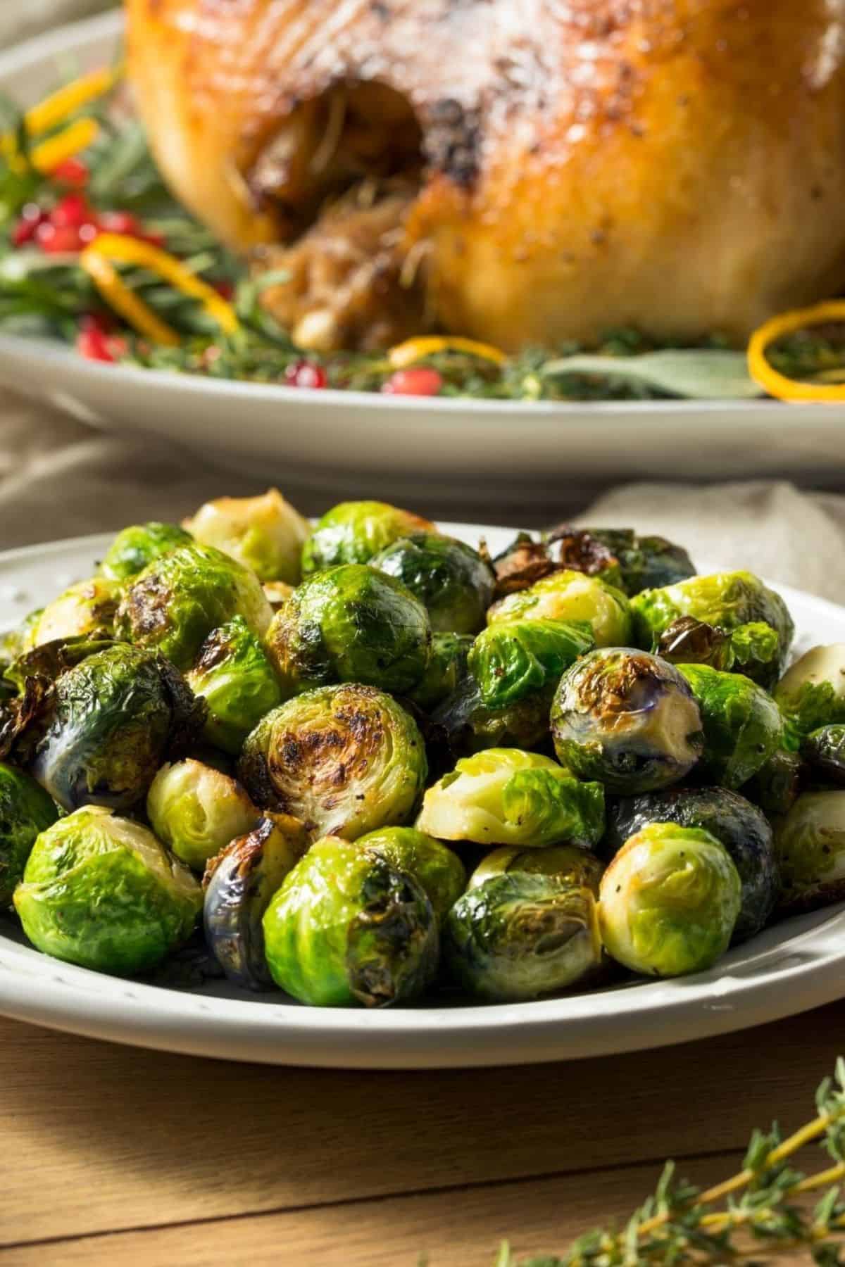 A plate of Roasted Brussels Sprouts with Parmesan Cheese sits in the foreground, with a cooked whole turkey garnished with herbs and citrus slices in the background.