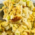 A close-up of a spoonful of Sourdough Stuffing with chopped celery and onions, held above a dish filled with more savory stuffing.