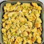 Square dish filled with featuring cubed bread, chopped celery, onions, and herbs, placed on a striped white and gray kitchen towel.