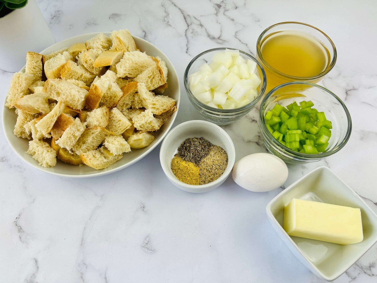 A bowl of cubed sourdough bread, chopped onion, celery, a bowl of broth, spices, a whole egg, and butter set on marble—everything you need to prepare delicious dish.