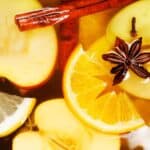 Close-up of apple slices, orange wedges, star anise, and a cinnamon stick floating in spiced drink.
