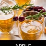 Three glasses of boilo, a warm amber drink, are garnished with rosemary sprigs and cranberries on a light table.