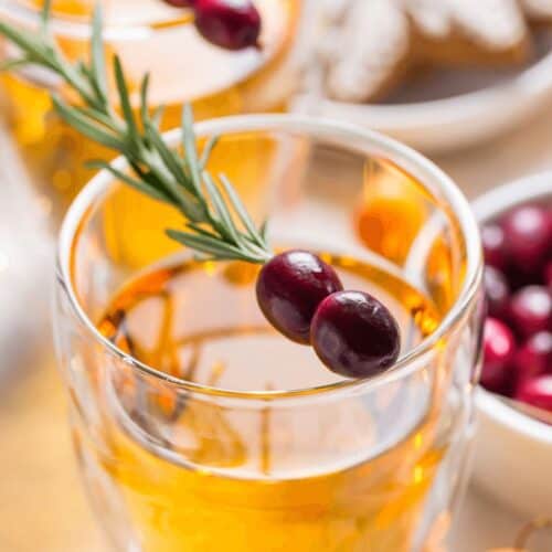 Two glasses of amber-colored boilo garnished with rosemary and cranberries, with cranberries and cookies visible in the background.
