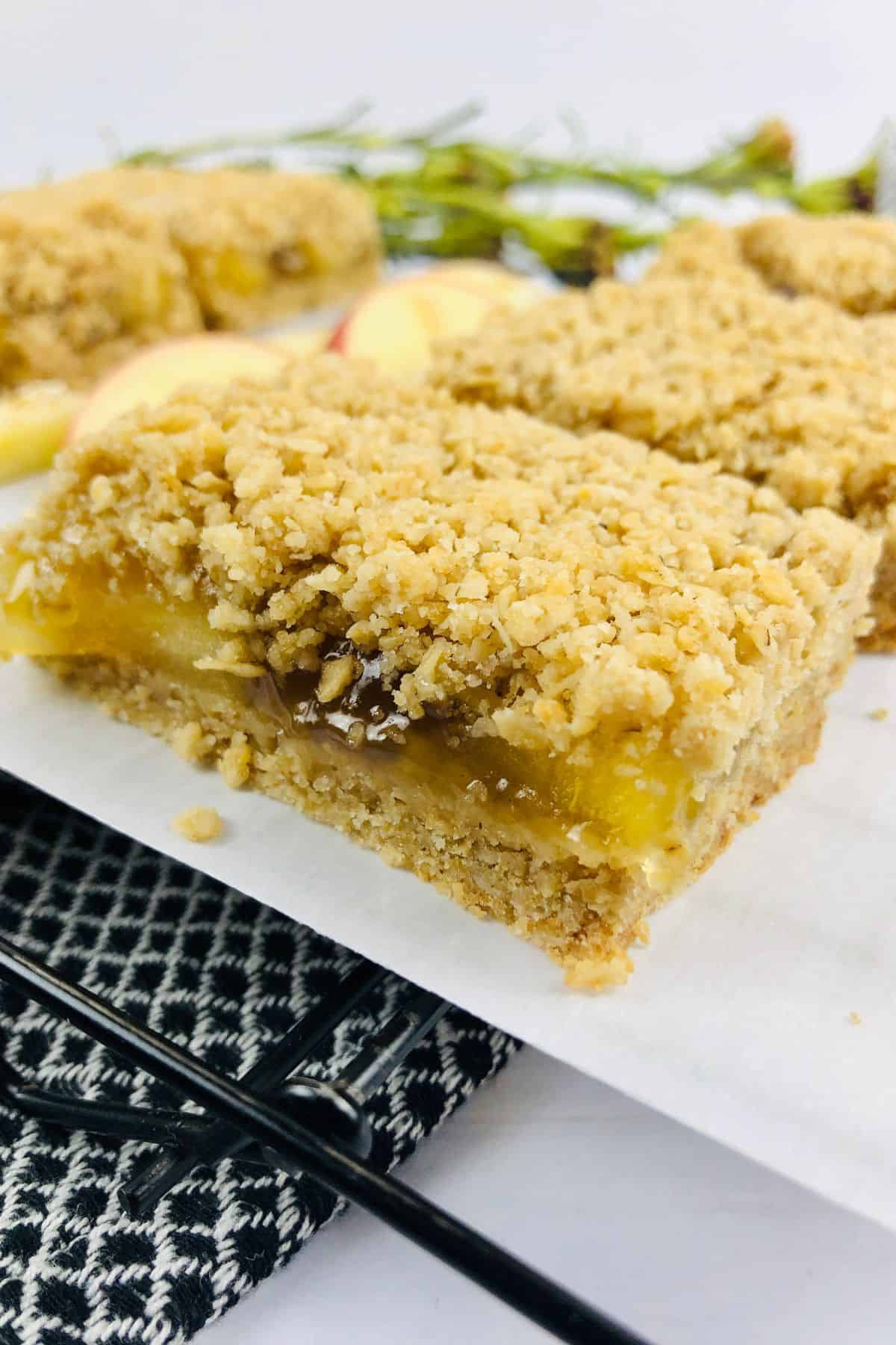 A close-up of a crumbly, layered apple oatmeal bar on parchment paper, with fresh apple slices and another apple oatmeal bar in the background.