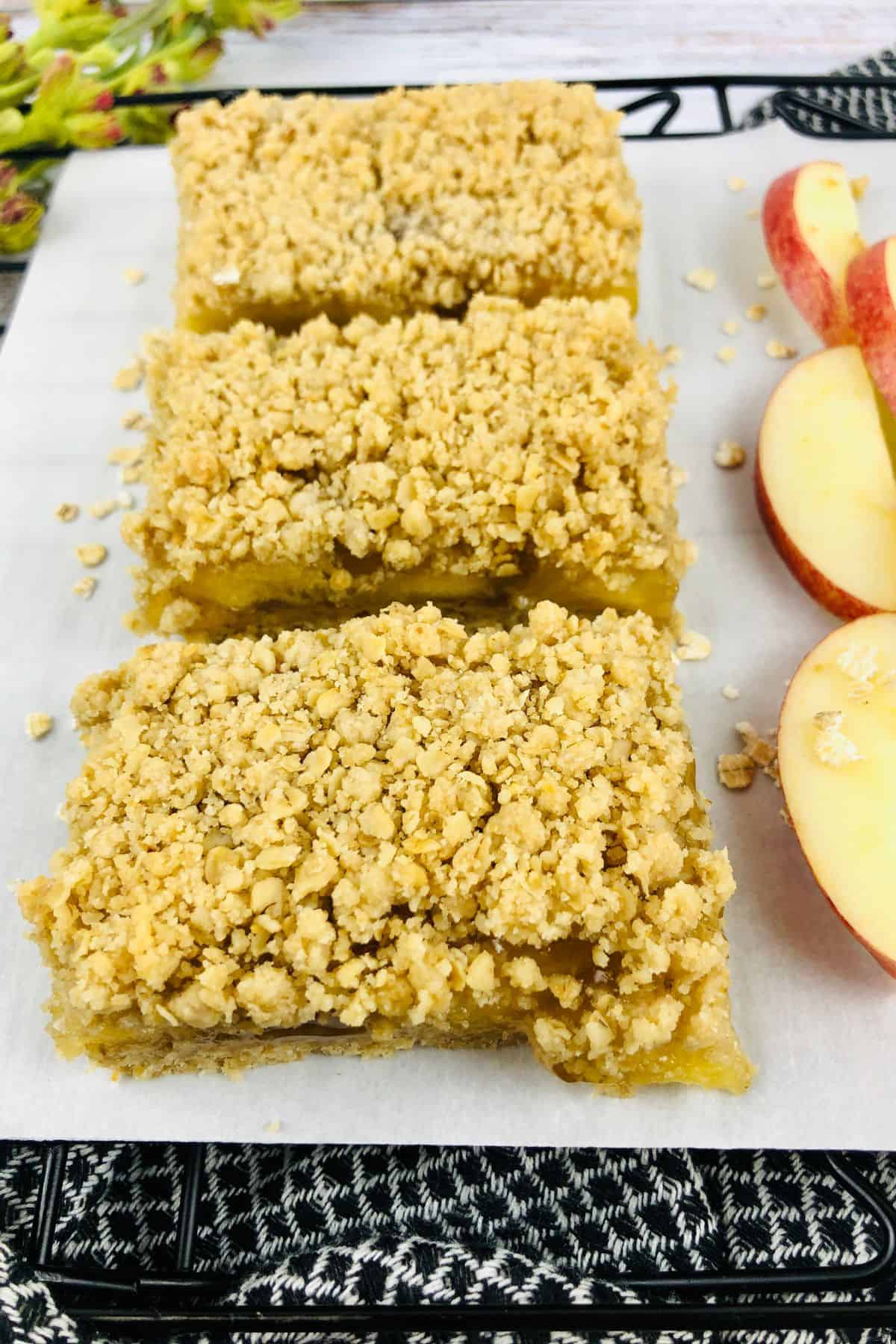 Three rectangular treats are arranged in a row on parchment paper next to several slices of fruit.