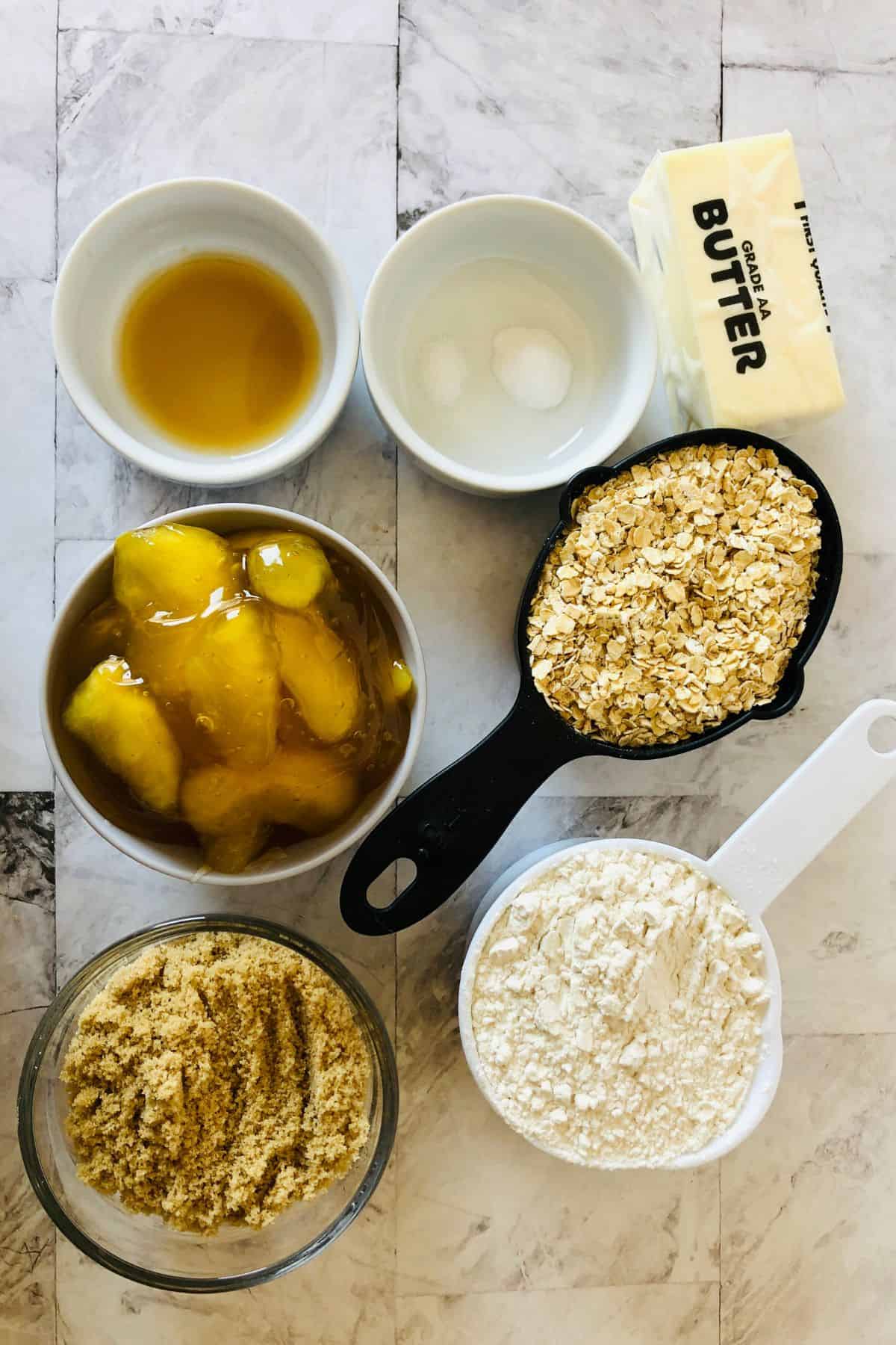 Ingredients for baking apple oatmeal bars are arranged on a countertop, including butter, oats, flour, brown sugar, apple filling, vanilla extract, and salt in measuring cups and bowls.