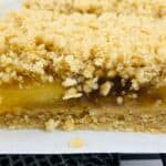 Close-up of a crumbly apple oatmeal bar with a visible fruit filling, resting on parchment paper and a patterned cloth.