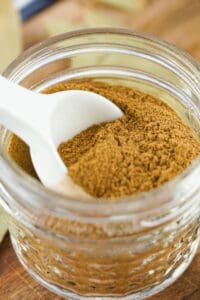 A glass jar filled with ground cinnamon and Apple Pie Spice, with a white spoon resting inside.