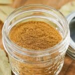 A small glass jar filled with ground brown Apple Pie Spice sits on a wooden surface, with a lid nearby.