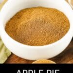 A white bowl filled with aromatic Apple Pie Spice sits on a wooden surface, with a bay leaf nearby and a small portion of spice in a measuring spoon.