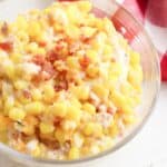 A bowl of creamy corn salad with bits of bacon sits invitingly on a white surface, reminiscent of a deconstructed corn casserole with cream cheese, beside a red and white checkered cloth.