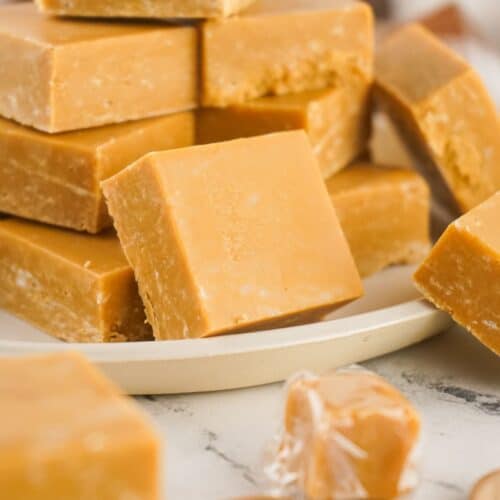 A plate stacked with square pieces of light brown caramel fudge, with a few pieces and one wrapped caramel in the foreground.