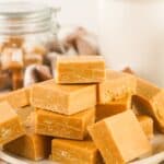 A plate stacked with several pieces of light brown caramel fudge, with jars and containers in the background.