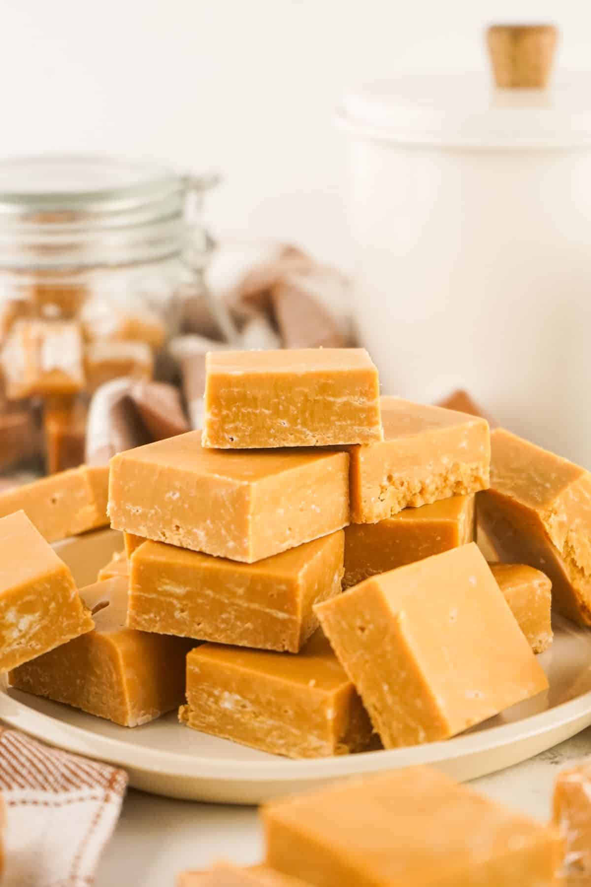 A plate stacked with several pieces of light brown caramel fudge, with jars and containers in the background.