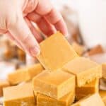A hand is picking up a square piece of caramel fudge from a plate stacked with similar light brown fudge pieces.