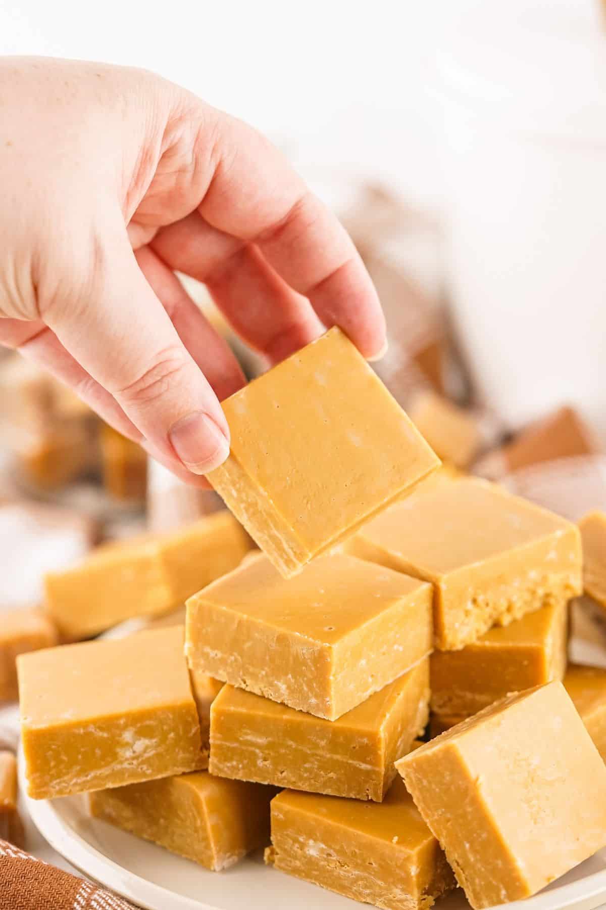 A hand is picking up a square piece of candy from a plate stacked with similar  pieces.