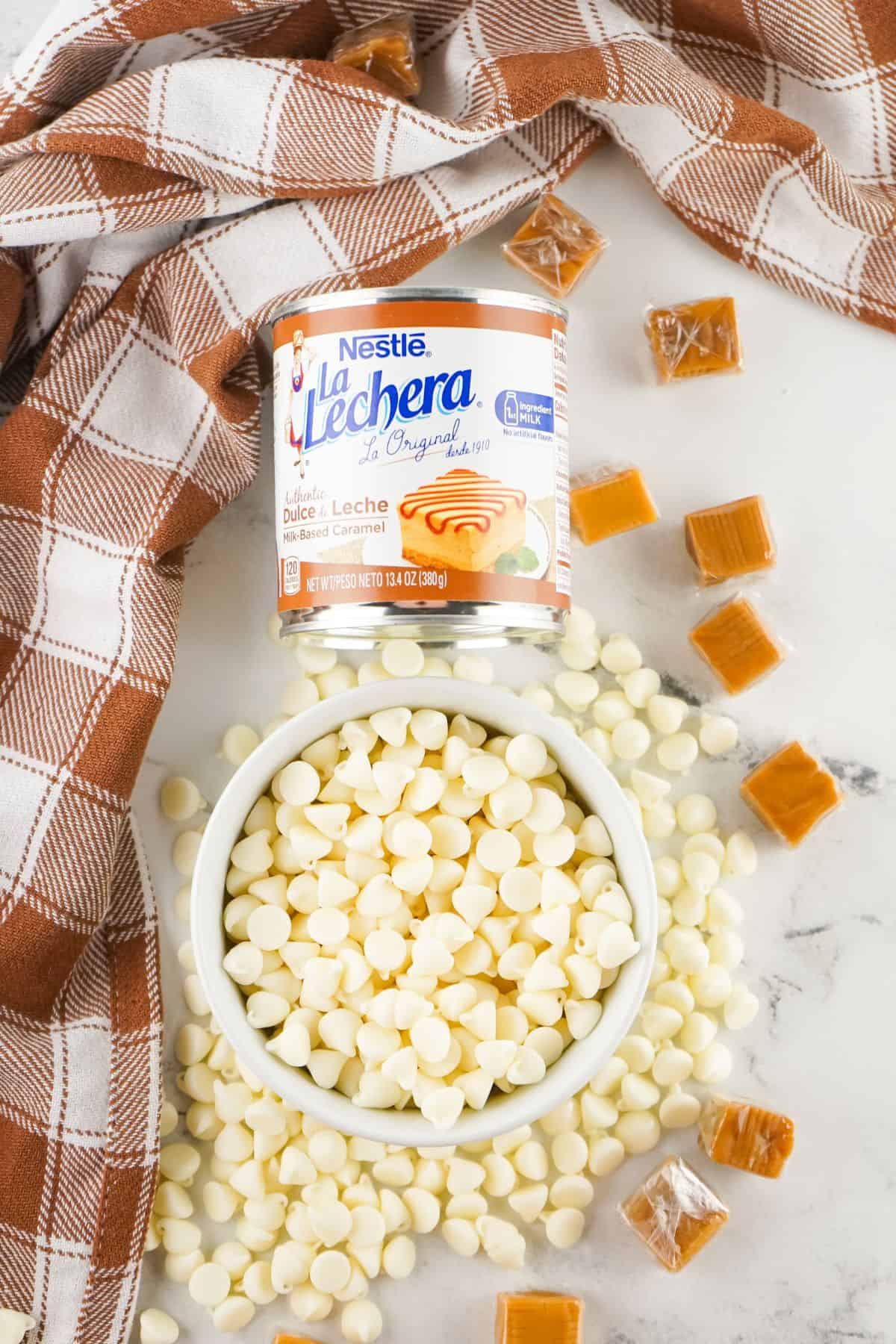 A can of Nestlé La Lechera dulce de leche sits beside a bowl of white chocolate chips and caramel fudge candies, all arranged on a white surface with a brown checkered cloth.