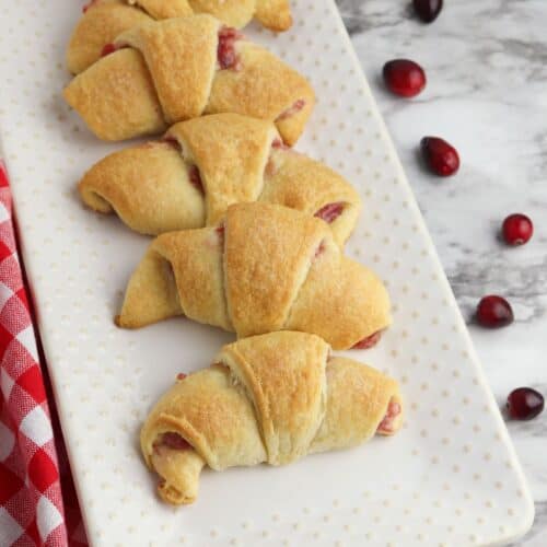 A rectangular white plate holds six golden brown Cheesecake Crescent Rolls with a red filling, placed on a marble surface with scattered cranberries and a red checked cloth.