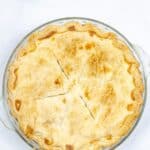 A golden, flaky crust, partially sliced into six pieces, sits in a clear glass pie dish on a white surface.