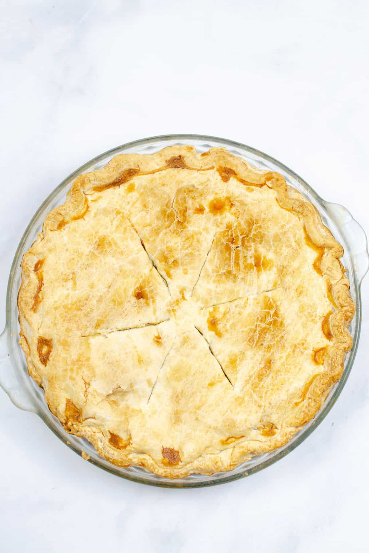 A golden, flaky crust, partially sliced into six pieces, sits in a clear glass pie dish on a white surface.