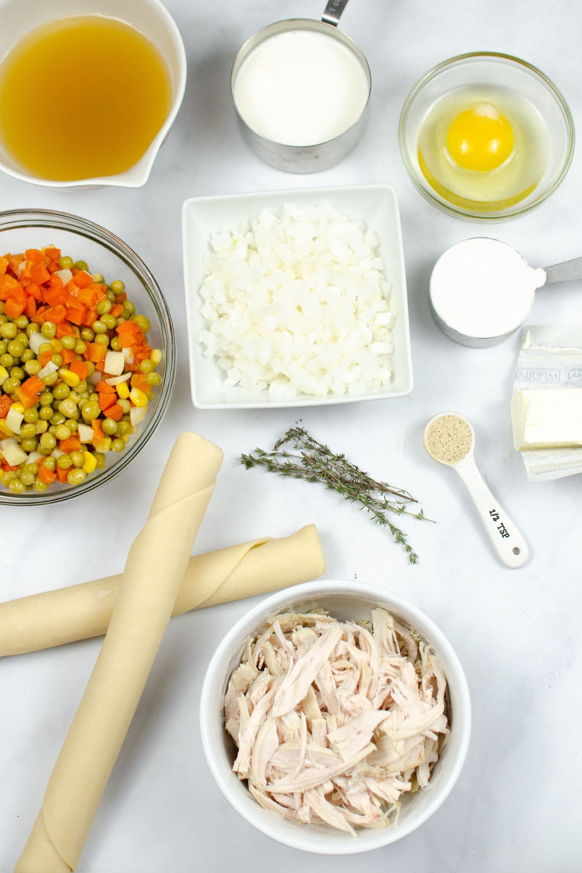 Ingredients arranged on a white surface, including pie crusts, shredded chicken or turkey, mixed vegetables, onion, broth, milk, egg, butter, flour, thyme, and salt.