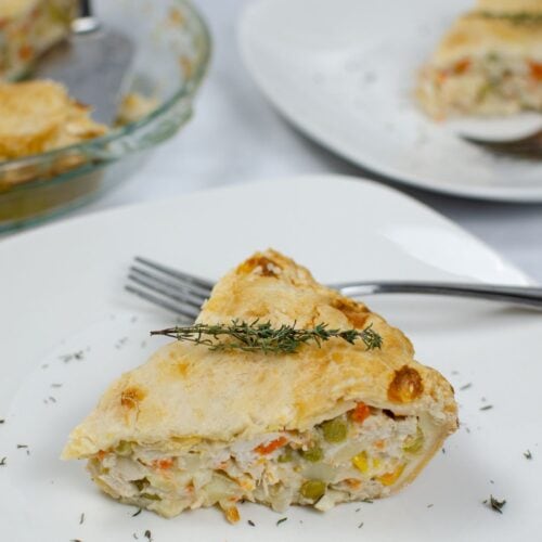 A slice of Easy Turkey Pot Pie with vegetables sits on a white plate, garnished with fresh thyme, a fork beside it and more pie in the background.