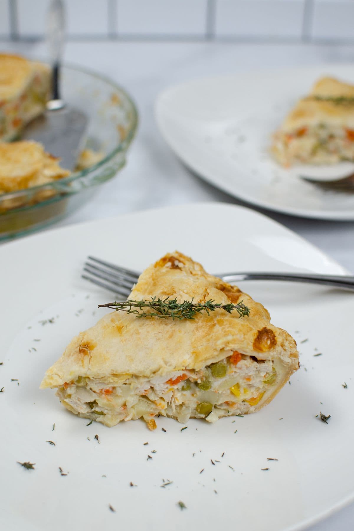 A slice of Easy Turkey Pot Pie with vegetables sits on a white plate, garnished with fresh thyme, a fork beside it and more pie in the background.