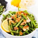 A white bowl of Green Beans and Almonds, garnished with toasted almond slices and a lemon wedge, sits on a blue cloth with sliced lemons and extra almonds in the background.