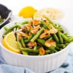 A white bowl filled with green beans and almonds, topped with toasted almond slices and garnished with a lemon wedge, sits on a blue cloth.