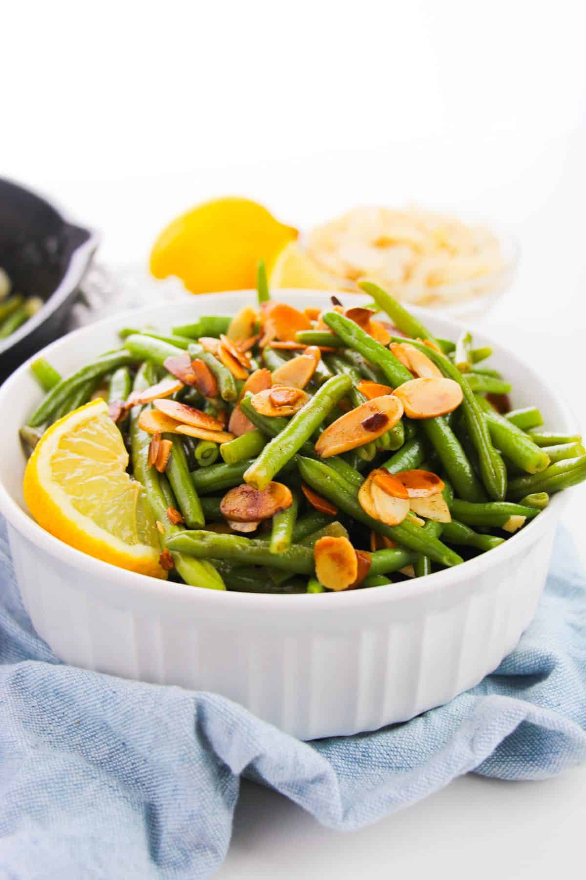 A white bowl filled with green beans and almonds, topped with toasted almond slices and garnished with a lemon wedge, sits on a blue cloth.