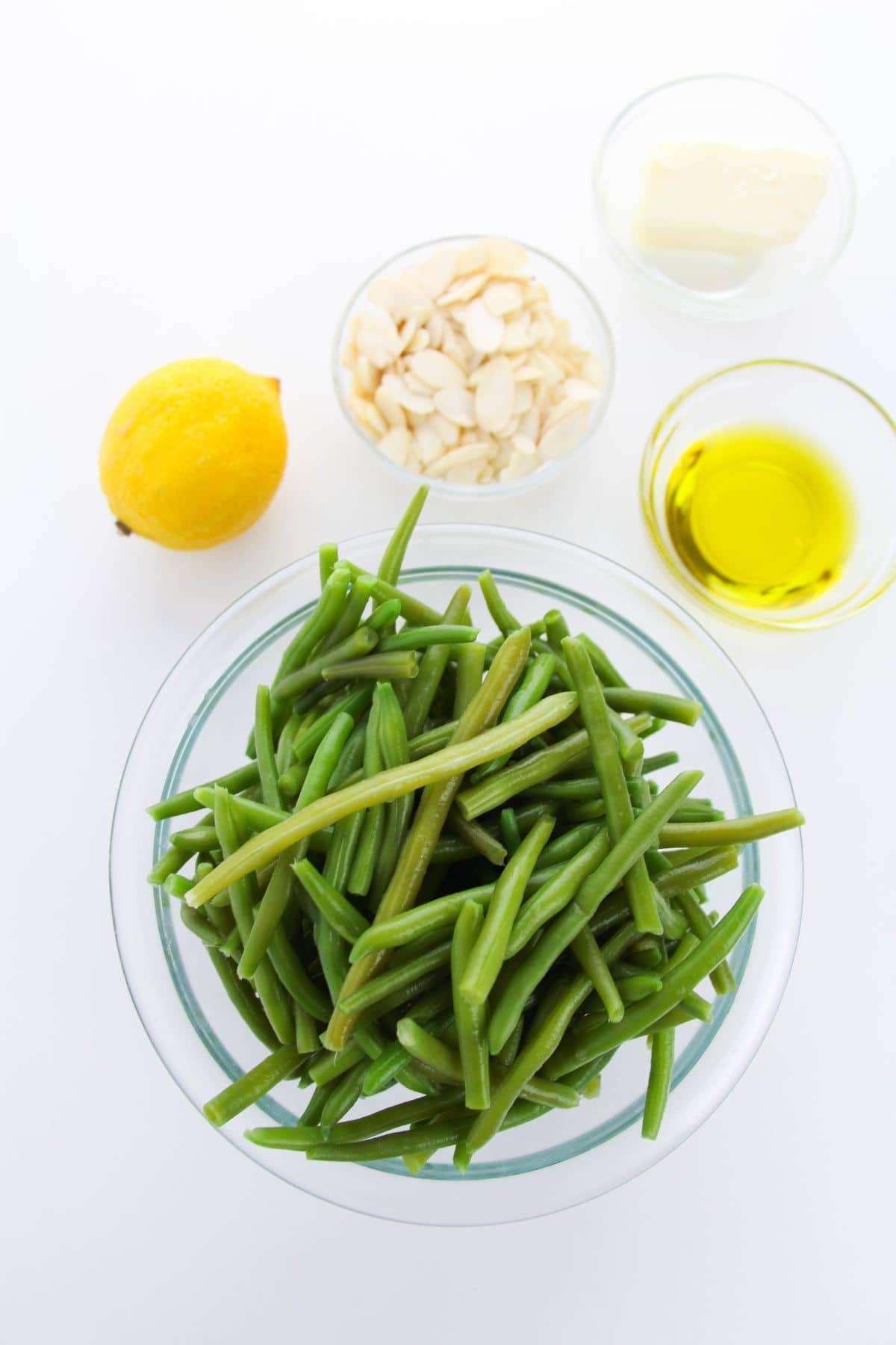 A bowl of fresh green beans and almonds is paired with lemon, butter, and olive oil in separate glass bowls on a white surface.