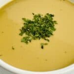 A bowl of creamy soup garnished with chopped fresh herbs and a drizzle of ham gravy, served in a white dish on a light surface.