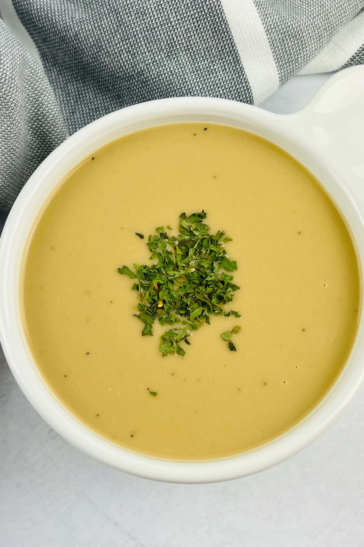 A bowl of creamy yellow soup topped with chopped green herbs, reminiscent of classic ham gravy, is placed next to a gray and white striped cloth.