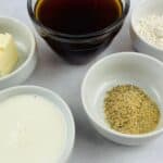 Five small bowls containing soy sauce, flour, butter, milk, and a mixture of salt and pepper are arranged on a white surface—perfect for preparing classic ham gravy.